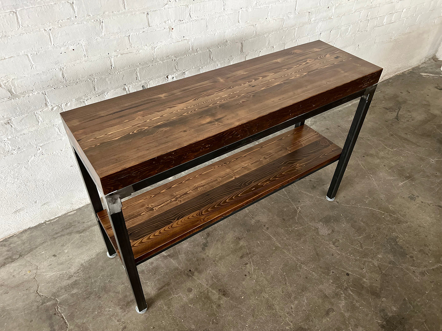 Grand Boulevard Console Table by Workshop Detroit - Made from reclaimed wood and steel - Shown in a walnut stain finish and sitting on a concrete floor with a white brick wall in the background