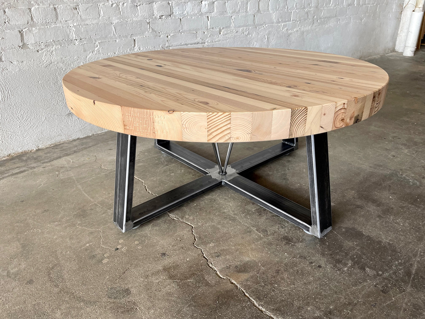 Grand Circus Coffee Table by Workshop Detroit - The coffee table has a round top made from reclaimed wood and a steel base - The coffee table is on a concrete floor with a white brick wall in the background