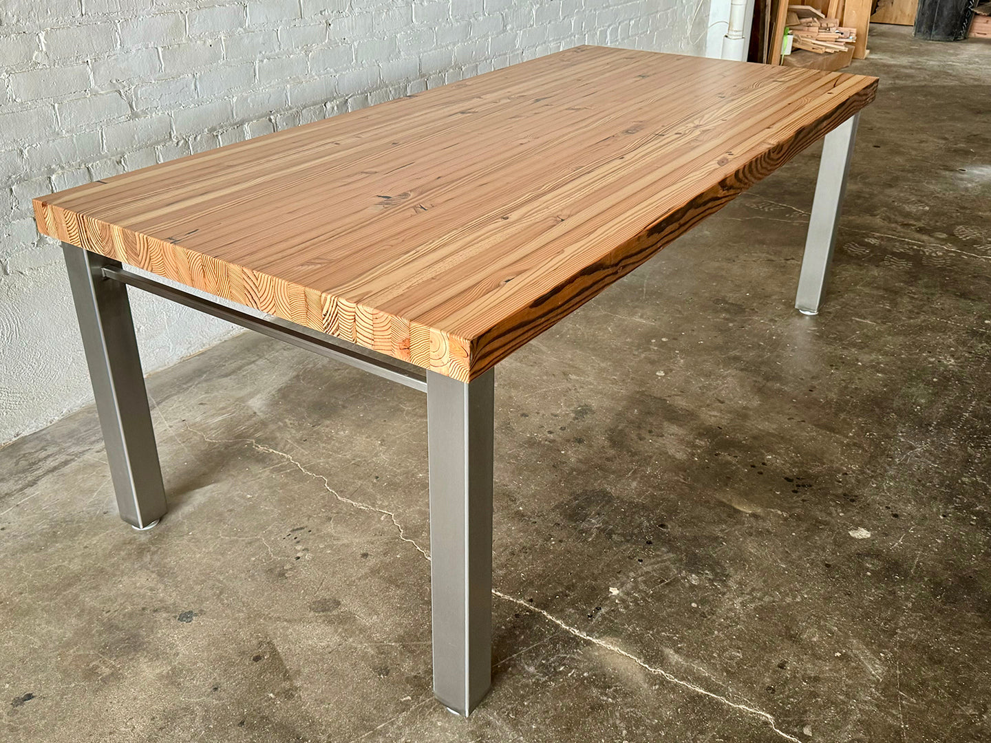 High Street Dining Table from Workshop Detroit - Made from solid reclaimed lumber with a stainless steel base - Shown in a natural clear coat finish on a concrete floor with a white brick wall in the background