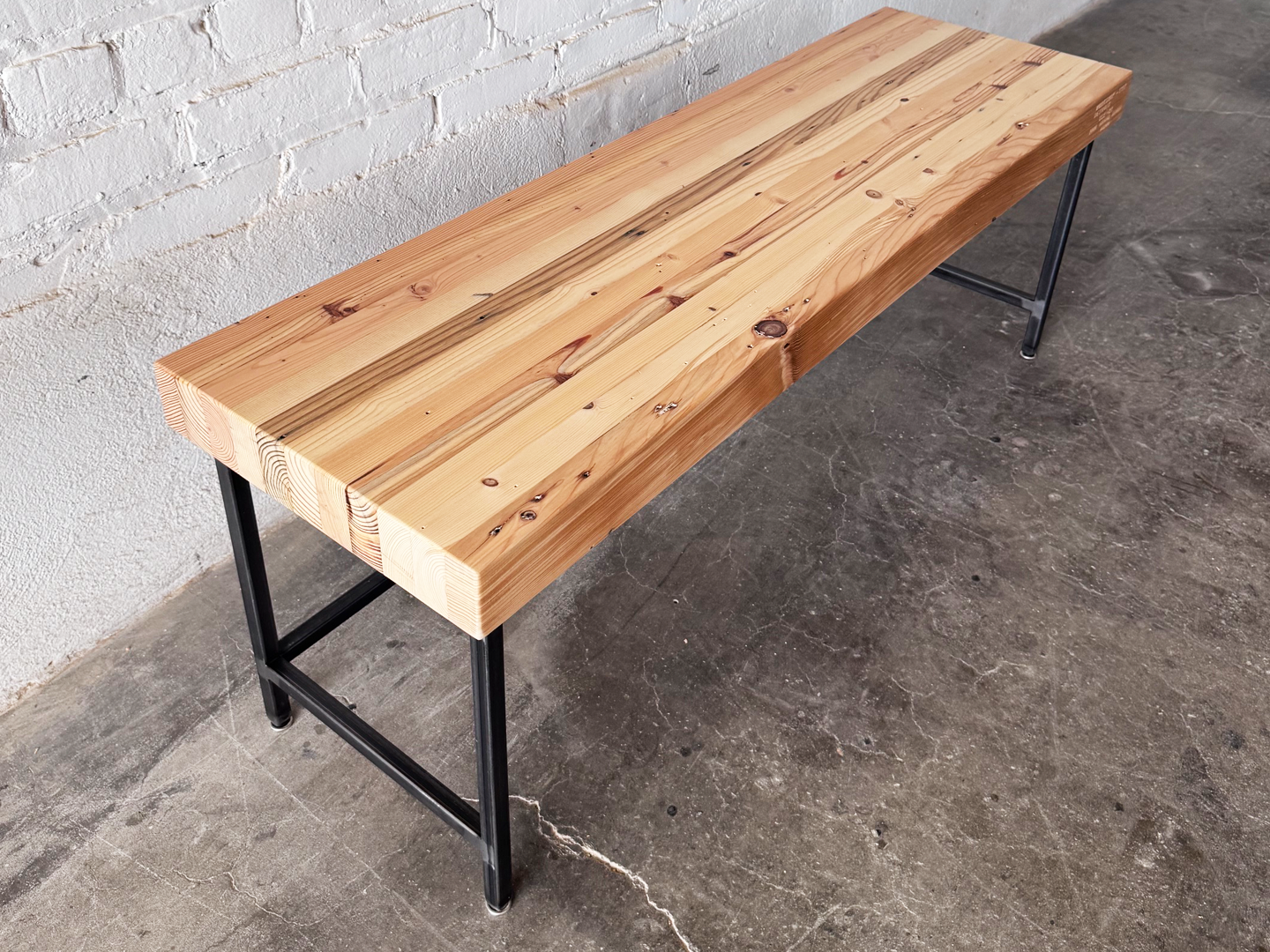 Top 3/4 view of the Grandy reclaimed wood and steel bench by Workshop Detroit shown next to a painted white brick wall and sitting on a concrete floor. Bench is shown with a clear, natural finish. 