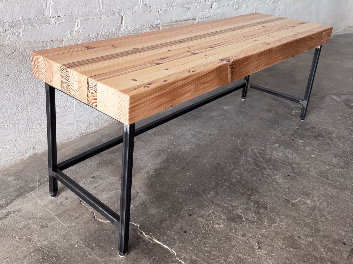 alternat view of the Grandy reclaimed wood and steel bench by Workshop Detroit shown next to a painted white brick wall and sitting on a concrete floor. Bench is shown with a clear, natural finish. 