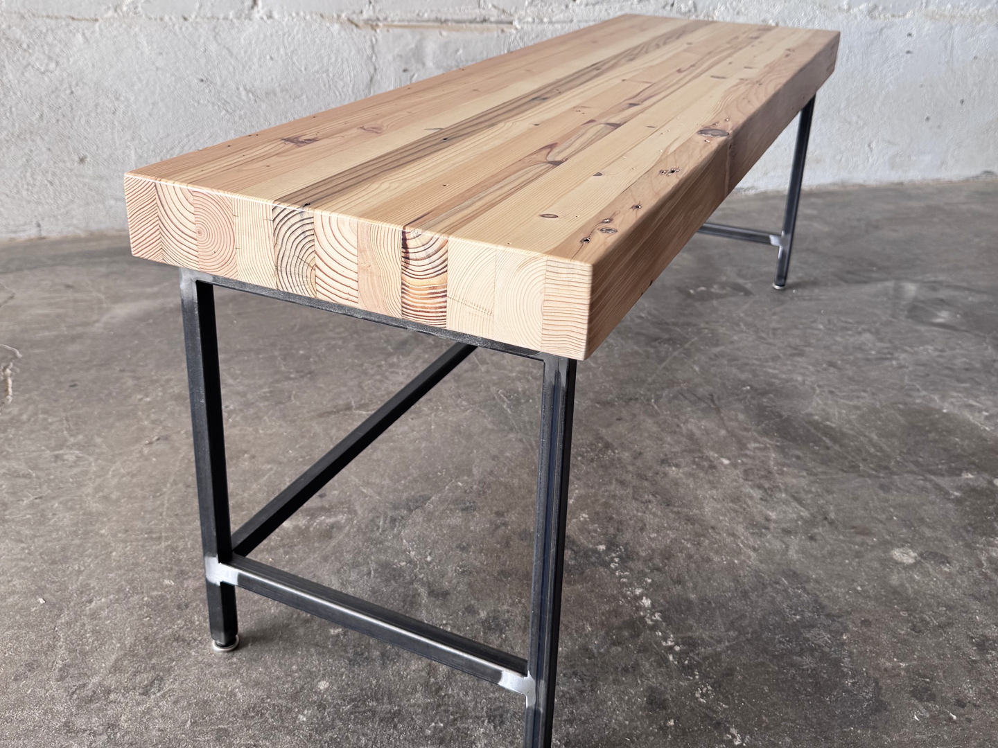 End 3/4 view of the Grandy reclaimed wood and steel bench by Workshop Detroit shown next to a painted white brick wall and sitting on a concrete floor. Bench is shown with a clear, natural finish. 