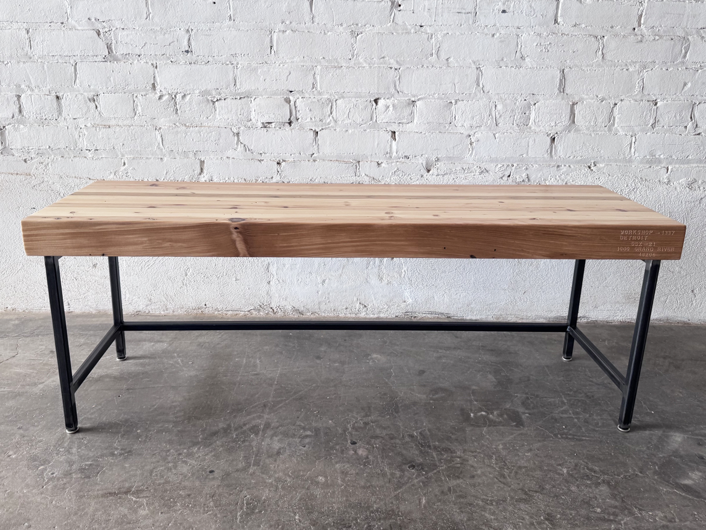 Side view of the Grandy reclaimed wood and steel bench by Workshop Detroit shown next to a painted white brick wall and sitting on a concrete floor. Bench is shown with a clear, natural finish. 