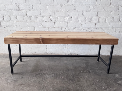 Side view of the Grandy reclaimed wood and steel bench by Workshop Detroit shown next to a painted white brick wall and sitting on a concrete floor. Bench is shown with a clear, natural finish. 