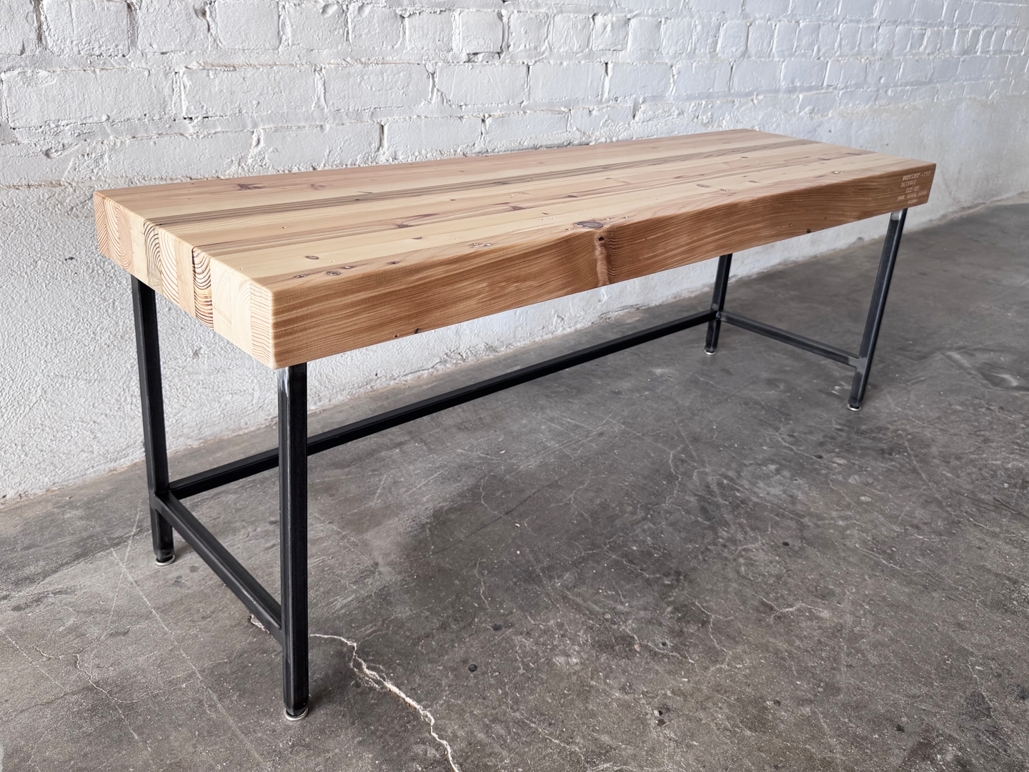 Grandy reclaimed wood and steel bench by Workshop Detroit shown next to a painted white brick wall and sitting on a concrete floor. Bench is shown with a clear, natural finish. 