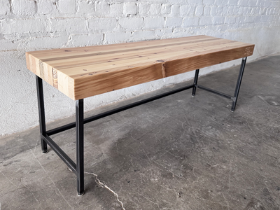 Grandy reclaimed wood and steel bench by Workshop Detroit shown next to a painted white brick wall and sitting on a concrete floor. Bench is shown with a clear, natural finish. 