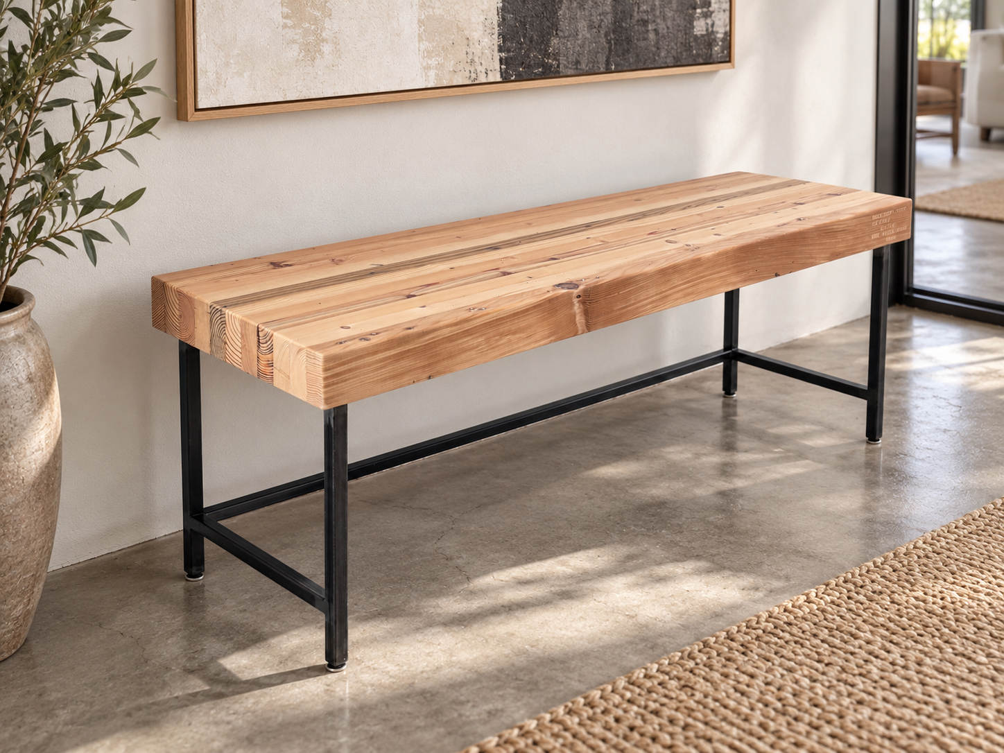 Grandy reclaimed wood and steel bench shown in a contemporary setting with white wall and concrete floor. Bench is shown with a clear, natural finish.