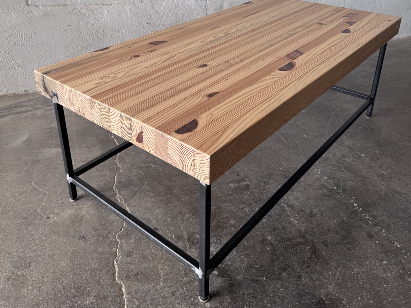 Alternate top 3/4 view of the Grandy Coffee Table by Workshop Detroit with a light natural wood top and a dark metal frame. Shown adjacent to a painted white brick wall and sitting on a concrete floor. 