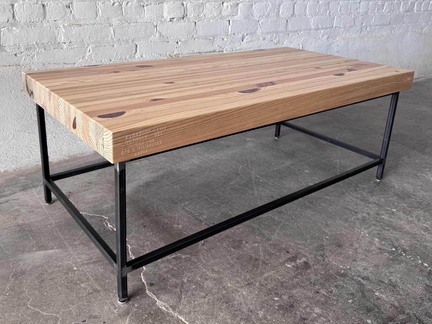 Alternate 3/4 mid view of the Grandy Coffee Table by Workshop Detroit with a light natural wood top and a dark metal frame. Shown adjacent to a painted white brick wall and sitting on a concrete floor. 