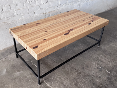 Top 3/4 view of the Grandy Coffee Table by Workshop Detroit with a light natural wood top and a dark metal frame. Shown adjacent to a painted white brick wall and sitting on a concrete floor. 