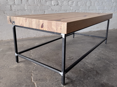 Low 3/4 view of the Grandy Coffee Table by Workshop Detroit with a light natural wood top and a dark metal frame. Shown adjacent to a painted white brick wall and sitting on a concrete floor. 