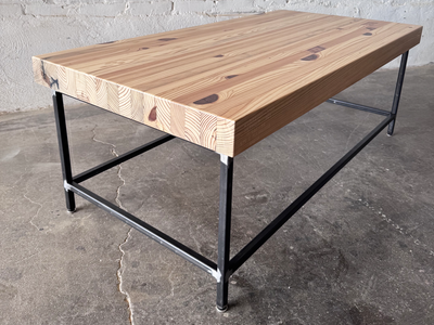 Grandy Coffee Table by Workshop Detroit with a light natural wood top and a dark metal frame. Shown adjacent to a painted white brick wall and sitting on a concrete floor. 