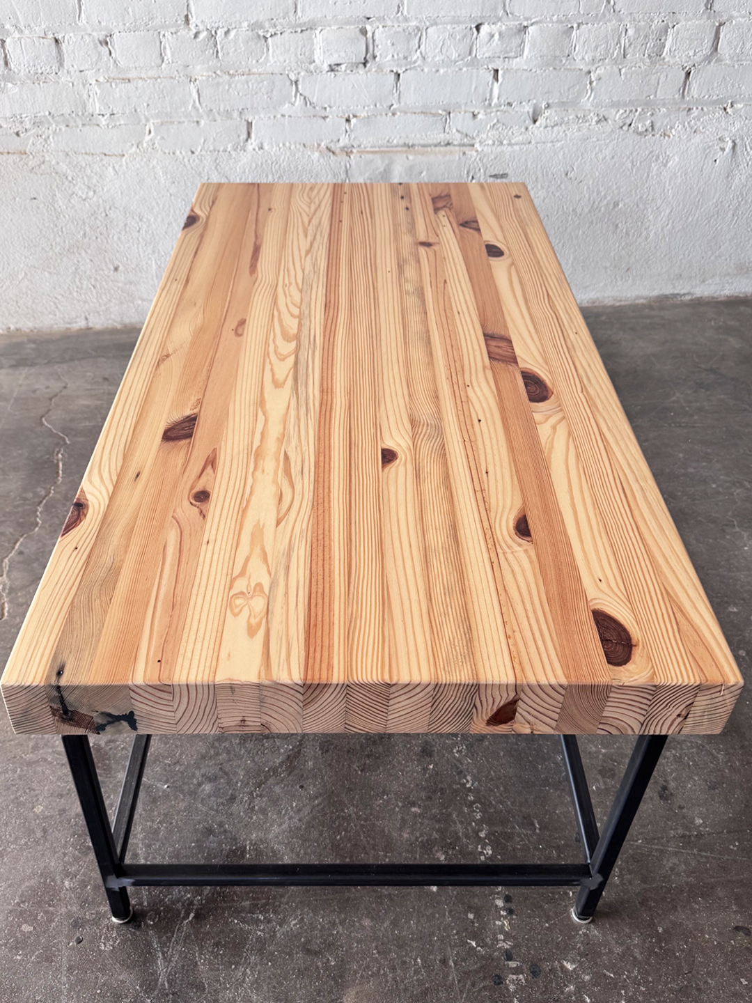 To end view of the Grandy Coffee Table by Workshop Detroit with a light natural wood top and a dark metal frame. Shown adjacent to a painted white brick wall and sitting on a concrete floor. 