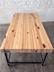 To end view of the Grandy Coffee Table by Workshop Detroit with a light natural wood top and a dark metal frame. Shown adjacent to a painted white brick wall and sitting on a concrete floor. 