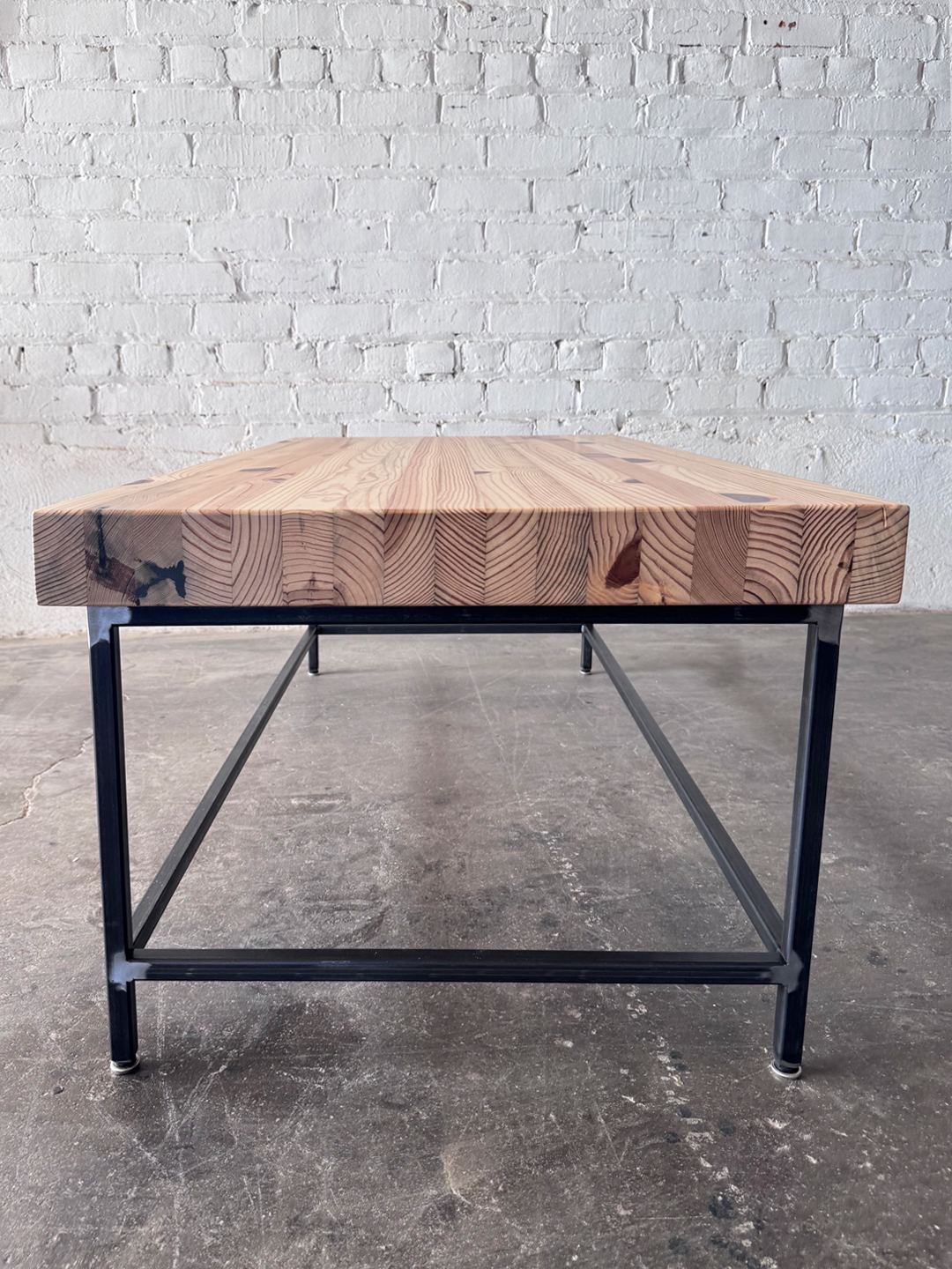 End view of the Grandy Coffee Table by Workshop Detroit with a light natural wood top and a dark metal frame. Shown adjacent to a painted white brick wall and sitting on a concrete floor. 