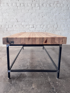End view of the Grandy Coffee Table by Workshop Detroit with a light natural wood top and a dark metal frame. Shown adjacent to a painted white brick wall and sitting on a concrete floor. 