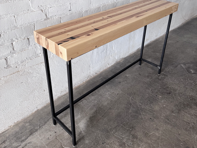 Alternate view of The Grandy Console Table with a light natural wood top and a black metal frame. Shown sitting on concrete floor next to a painted white brick wall. Shown with a natural finish.