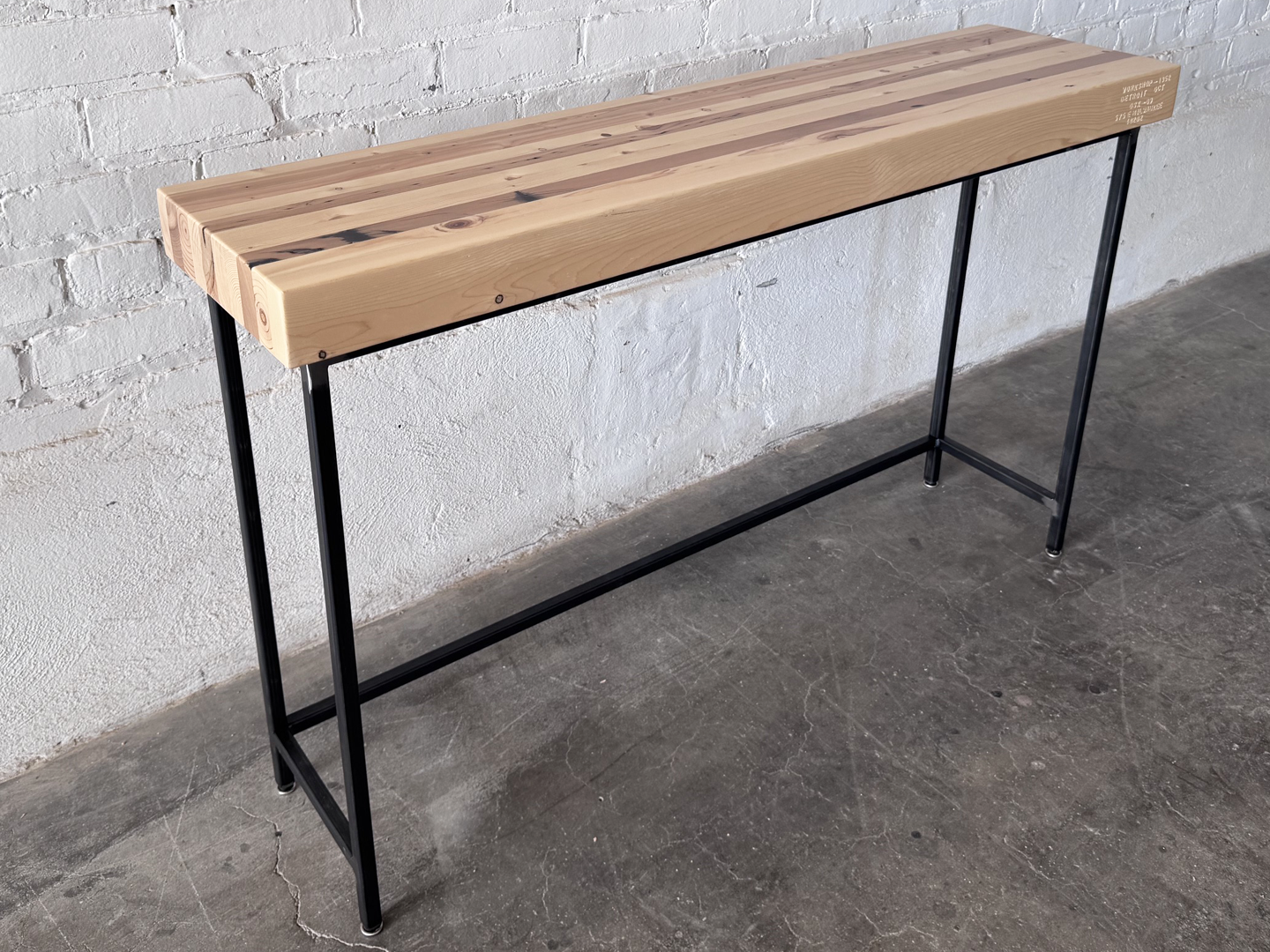 The Grandy Console Table with a light natural wood top and a black metal frame. Shown sitting on concrete floor next to a painted white brick wall. Shown with a natural finish.