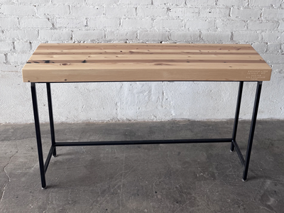 Side view long ways of the Grandy Console Table with a light natural wood top and a black metal frame. Shown sitting on concrete floor next to a painted white brick wall. Shown with a natural finish.