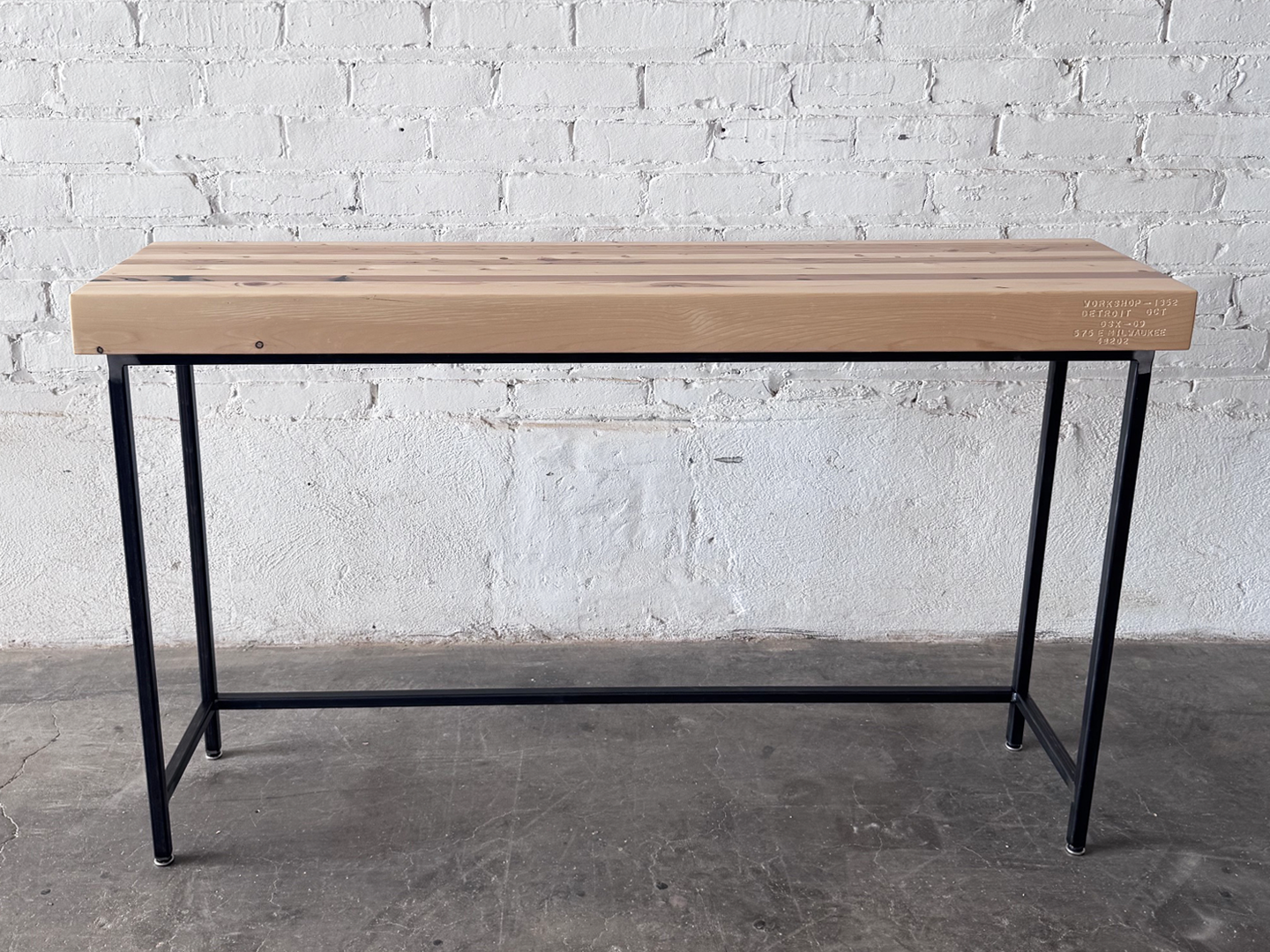 Alternate side view long ways the Grandy Console Table with a light natural wood top and a black metal frame. Shown sitting on concrete floor next to a painted white brick wall. Shown with a natural finish.