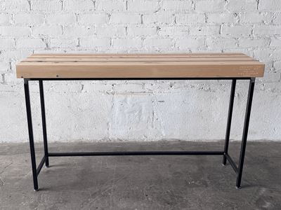 Alternate side view long ways the Grandy Console Table with a light natural wood top and a black metal frame. Shown sitting on concrete floor next to a painted white brick wall. Shown with a natural finish.
