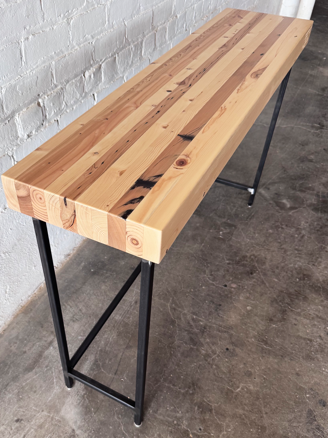 Top 3/4 view of The Grandy Console Table with a light natural wood top and a black metal frame. Shown sitting on concrete floor next to a painted white brick wall. Shown with a natural finish.