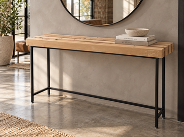 The Grandy Console Table with a light natural wood top and a black metal frame, styled in a contemporary entryway with a round mirror, books, and a bowl on top. Shown sitting on concrete floor. Shown with a natural finish.