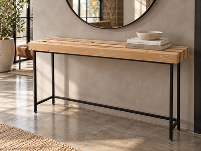 The Grandy Console Table with a light natural wood top and a black metal frame, styled in a contemporary entryway with a round mirror, books, and a bowl on top. Shown sitting on concrete floor. Shown with a natural finish.